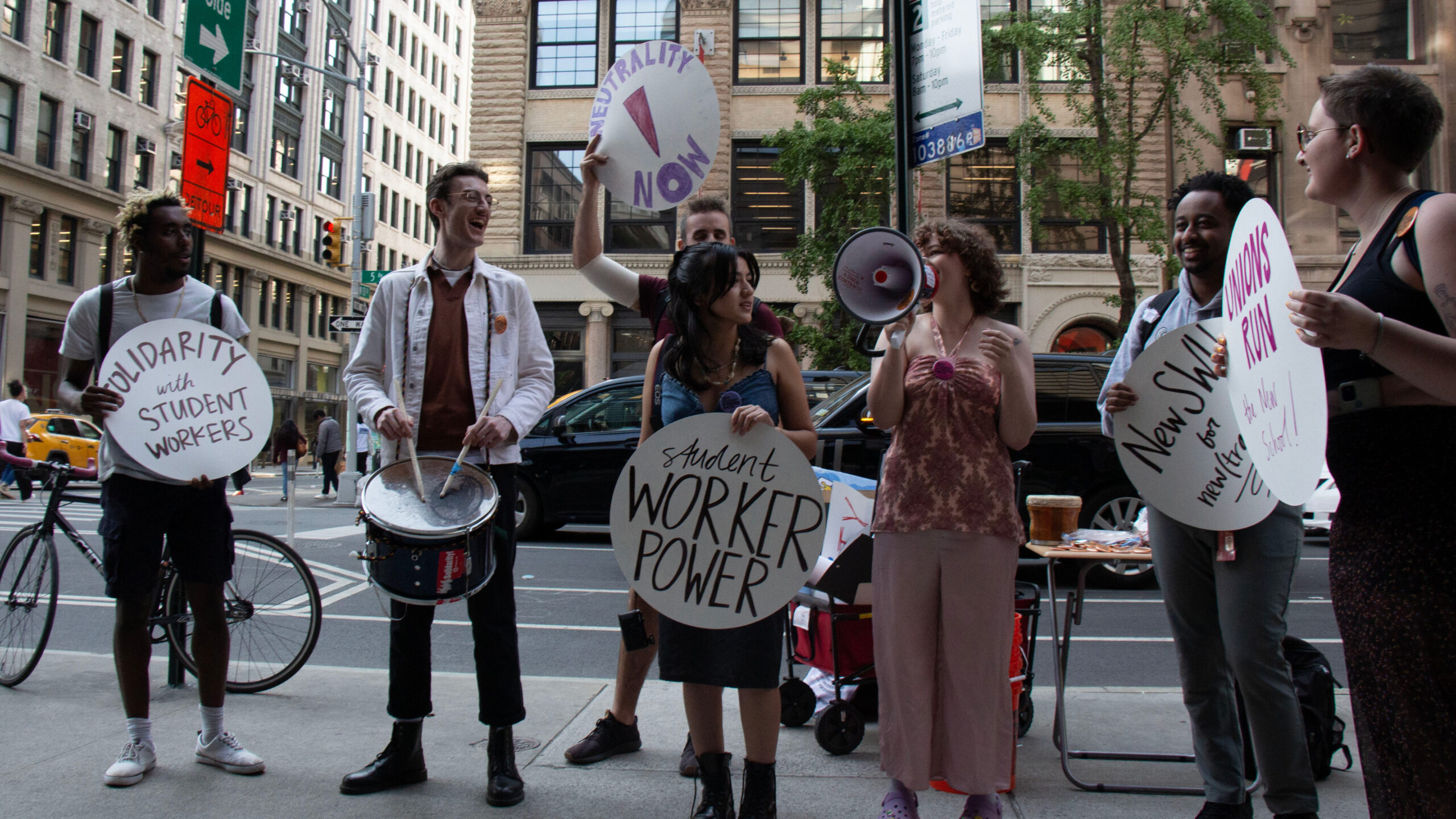 Seven students standing on the sidewalk during a rally. Five of them hold protest signs that read “Solidarity with Student Workers”, “Neutrality Now”, “Student Worker Power”, “NewSWU for Neutrality”, and “Unions Run the New School”. One student holds a drum and one holds a megaphone.