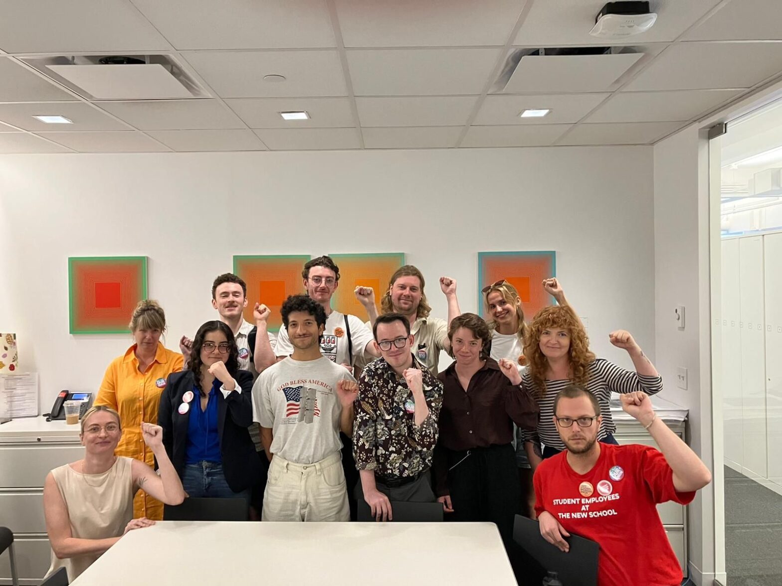 12 people have their fists raised as they face a camera. 10 stand in the middle of the group in two rows; one person sits on the left and on the right side. Behind them are three orange paintings hung on a white wall. In front of them is a white table.