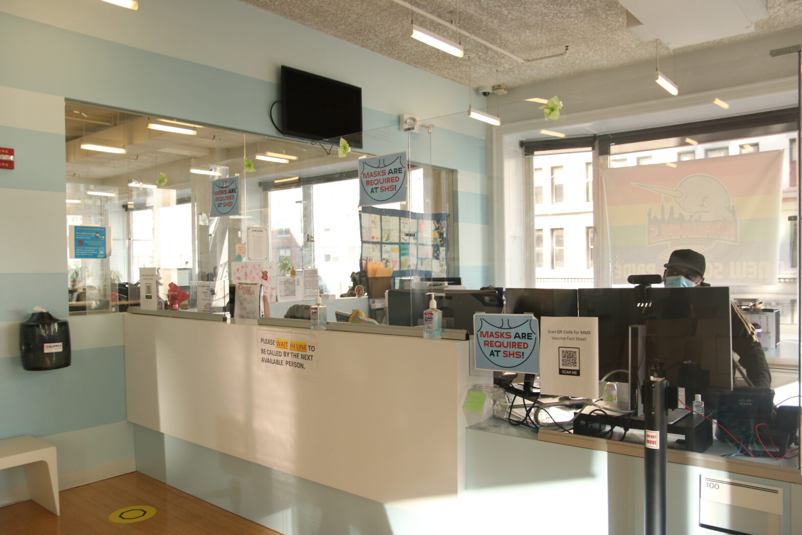 A large white and light blue desk with glass barriers is in a room with striped walls of the same colors and a large glass window behind the desk. The desk and glass have three signs that read “masks are required at SHS.” and one sign that reads “please wait in line to be called by the next person.” A Black man with a blue face mask sits behind a computer at the desk.