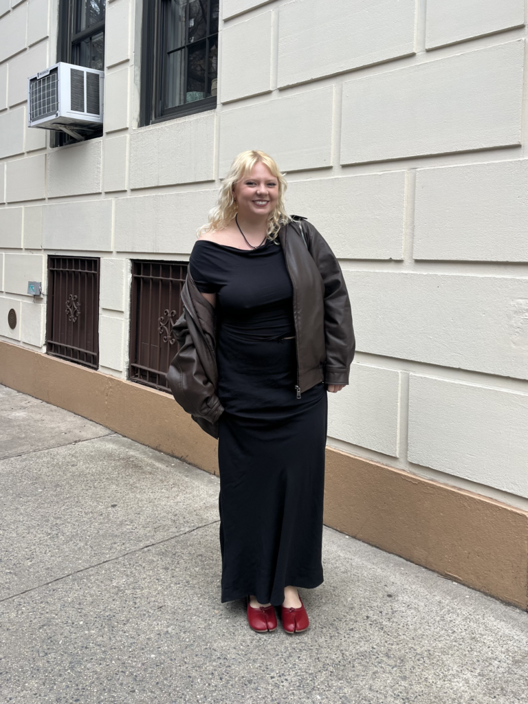 Student stands in front of a beige building wearing a brown leather jacket, black top, black maxi skirt, and red tabi ballet flats.