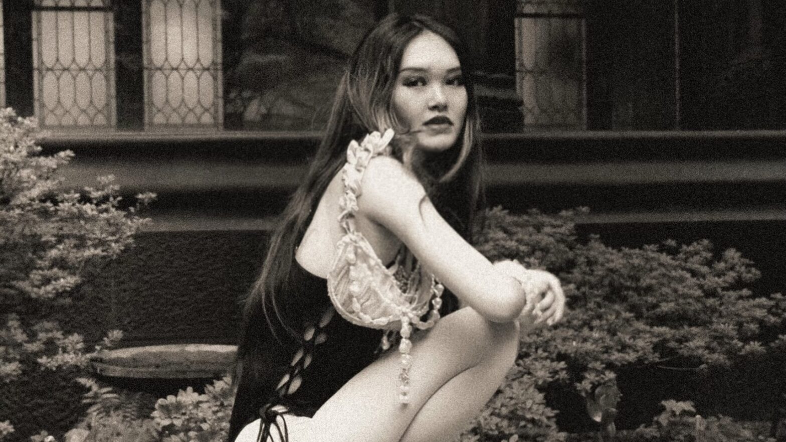 Black-and-white photo of a kneeling woman dressed in a black lace-up leotard, Mary Jane heels, and a small shoulder bag with dangling pearls and a ruffled strap.