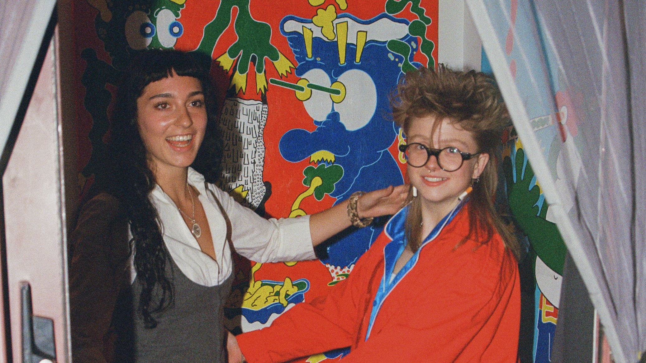 From left to right, one girl with long black hair and one girl with long blonde hair and black circular glasses smile in front of a red, blue, green, and yellow painting.