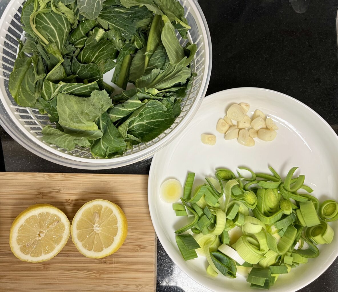 A bowl of chopped kale, a plate with chopped garlic and leek, and a cutting board with a lemon cut in half sit on a black counter.