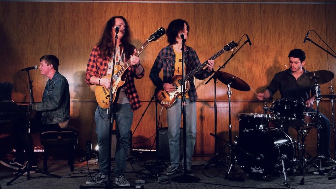 Four students play guitar, piano, and drums with microphones against a wood wall.