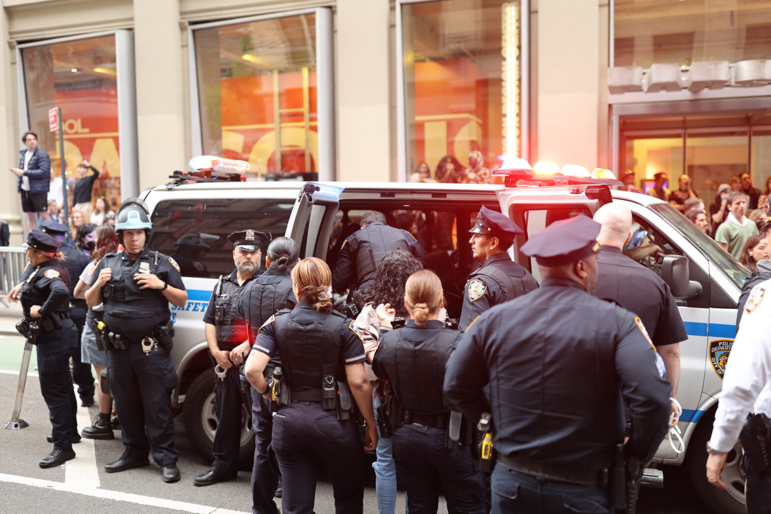 Several police officers lead an arrested individual into a New York Police Department vehicle in front of the 2 W. 13th Street Parsons building. Behind the vehicle, there are onlookers and demonstrators.