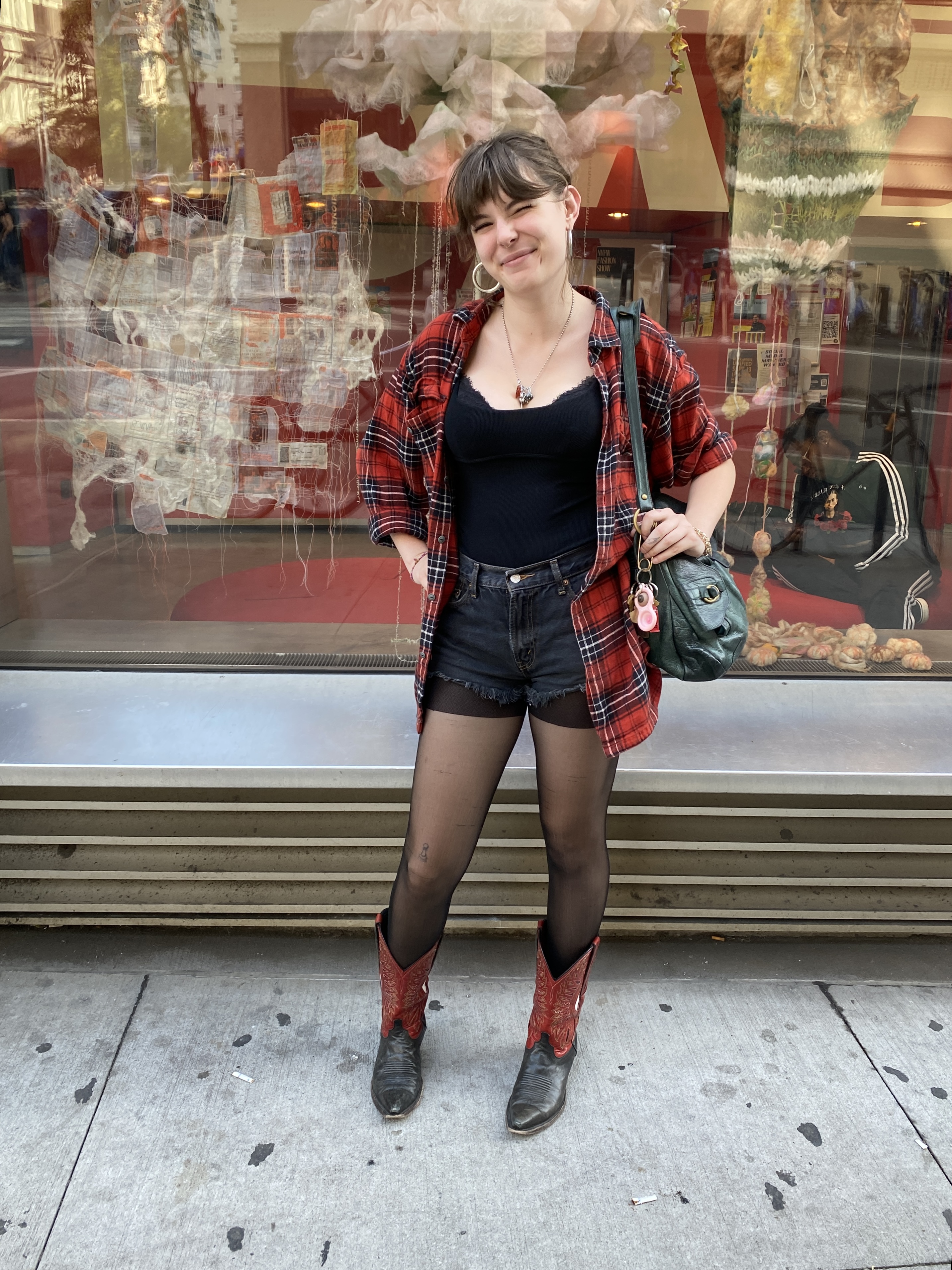 Student stands in front of the Parsons 13th Street building wearing a plaid black and red flannel ove ra black tank top, dark wash denim shorts, tights, and red and black cowboy boots, while carrying a green purse.