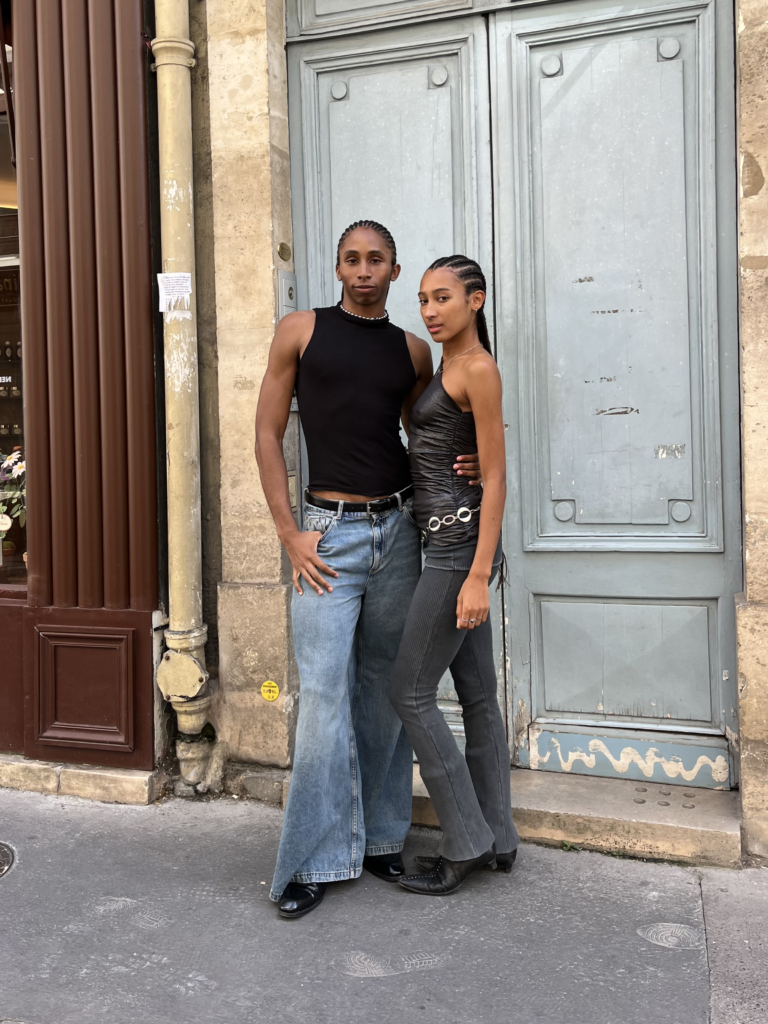 Two students wearing a black tank top, blue jeans, black boots, and a shiny black tank top, silver belt, gray pants, and black pointed boots stand in front of an old blue door in Paris, France.