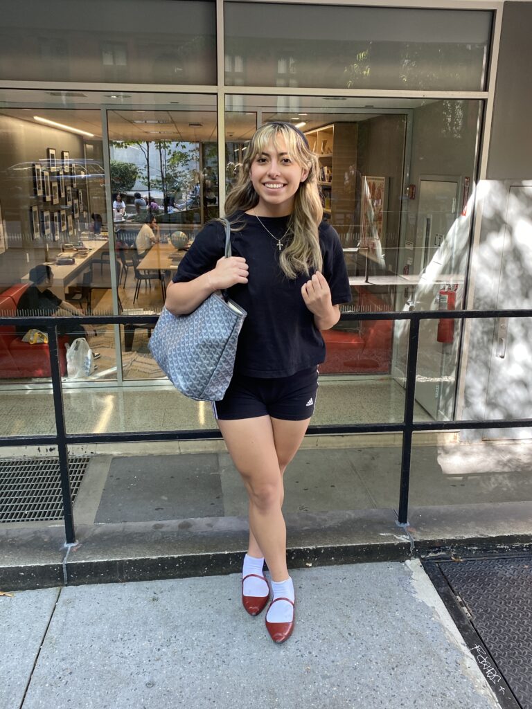 Student stands in front of the Lang 11th Street building wearing a black T-shirt, black Adidas biker shorts, white socks, a cross necklace, and red Mary Janes while carrying a gray, abstract print purse.