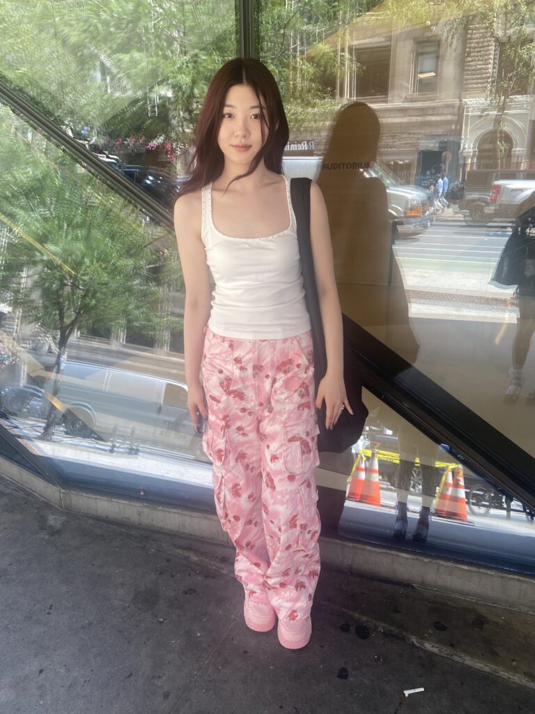 Student stands in front of the University Center wearing a white tank top, pink cargo pants with strawberry print, pink sneakers, and carries a black bag on their shoulder.