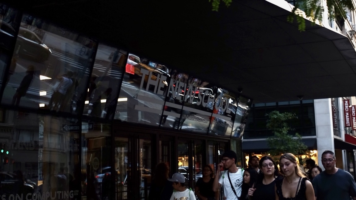 With the camera looking slightly upward, the front doors and awning of The New Nchool’s University Center are pictured with a few branches of a curbside tree in the foreground. A crowd of walking New Yorkers are featured in the lower right-hand corner.