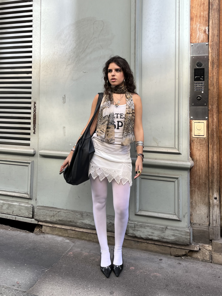 A student wearing a white tank top, white skirt, white tights, leopard print scarf, and black kitten heels that match her black tote bag stands in front of a blue wall in Paris, France.