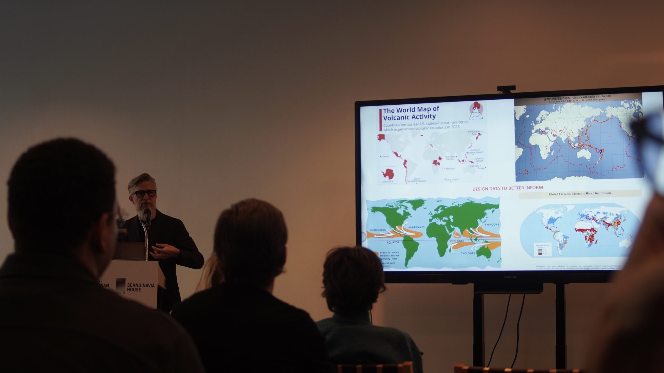 Man with facial hair talking behind a podium next to a TV screen showing a global map of areas with volcanic activity.