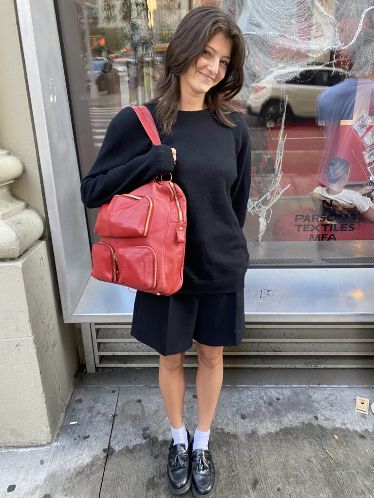 Student stands in front of the Parsons 13th Street building wearing a black sweater, black shorts, white socks, black loafers, and carrying a red bag with three pockets.