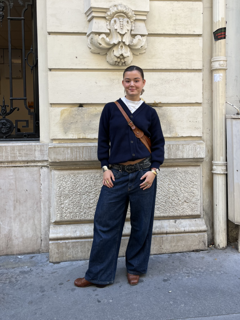 A student wearing a navy blue cardigan, white tshirt, navy blue jeans, and a brown leather crossbody bag that matches her brown leather boots stands in front of an ornamental beige stone wall in Paris, France.