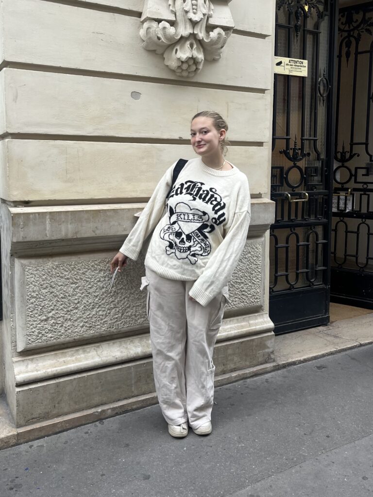 Student wearing a white knit Ed Hardy sweater, white cargo pants, and white sneakers stands against a beige building in Paris, France.