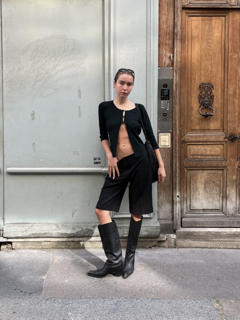 Student wearing a black cardigan, black knee-length shorts, and black knee-high boots stands against a light blue wall and wooden door in Paris, France.