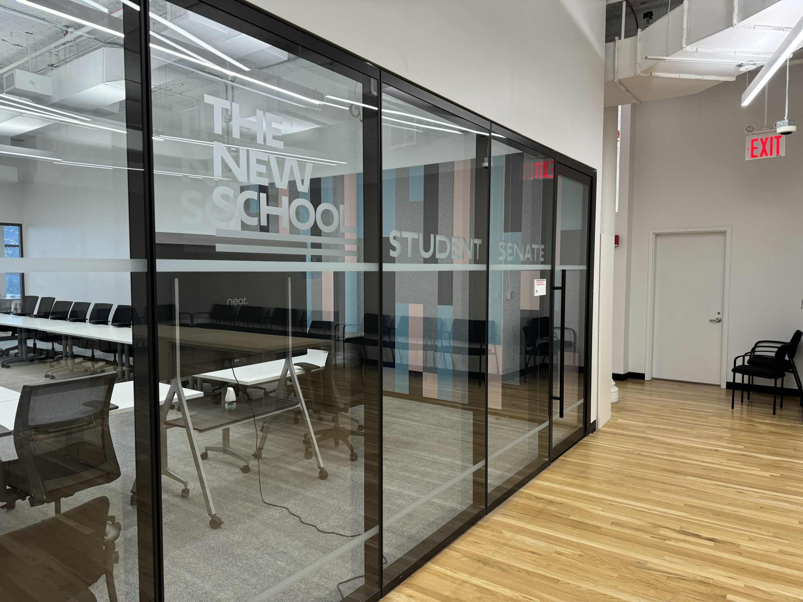 A glass room with the letters “New School Student Senate” written in gray, all capital letters is photographed on an angle. Inside the room are white tables in a rectangle with black chairs surrounding them and a television in front. Outside the room is a light brown hardwood floor; further back is a white wall, white door, and two black chairs.