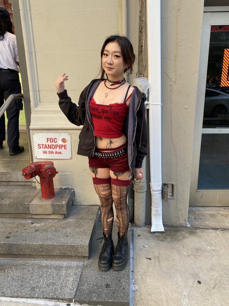 Student stands in front of the Parsons 13th Street building wearing a cropped red tank top, red micro shorts, a black belt, a gray jacket, red and gold necklaces, red garters, ripped black tights, and black Doc Martens.