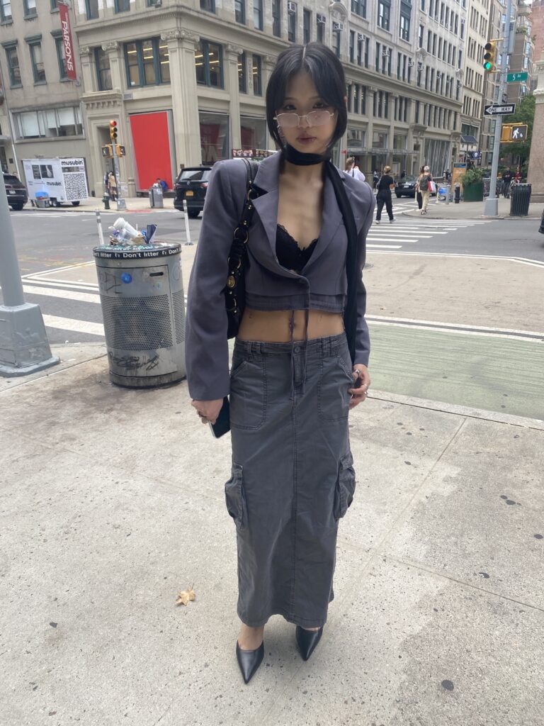 Student stands in front of the University Center wearing a cropped gray blazer, a black choker, a black lacy bra, a gray cargo maxi skirt, black heels, and carries a black handbag on their shoulder.