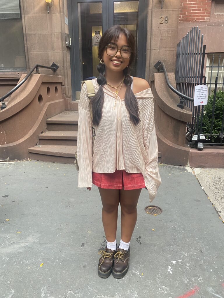 Student stands in front of the building across the street from the Lang 12th Street building wearing a peach off-the-shoulder long-sleeve top, a red denim skort, white crew socks, and brown shoes.