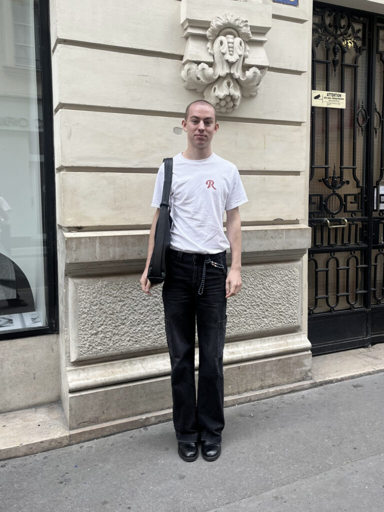 Student wearing a white t-shirt, black jeans, black boots, and a black bag stands against a beige building in Paris, France.