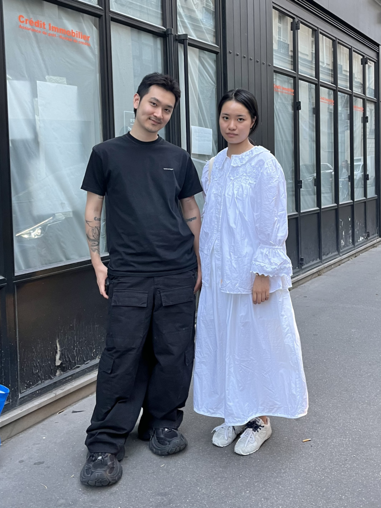 Two students, one wearing an all black Balenciaga outfit and the other wearing an all white outfit stand in front of a black storefront with glass windows in Paris, France.