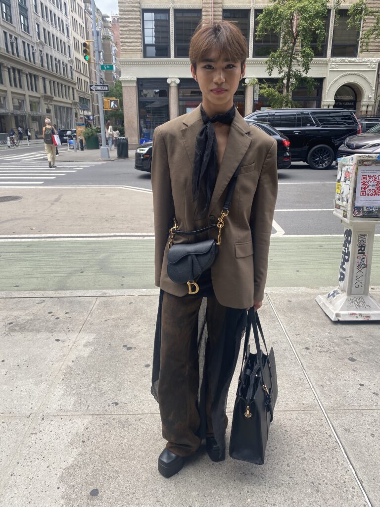 Student stands in front of Fifth Avenue wearing a brown blazer, a black scarf that ties around their neck and flows to their legs, brown wide-legged plants, black platform boots,and a black Dior crossbody bag, all while carrying a black handbag.