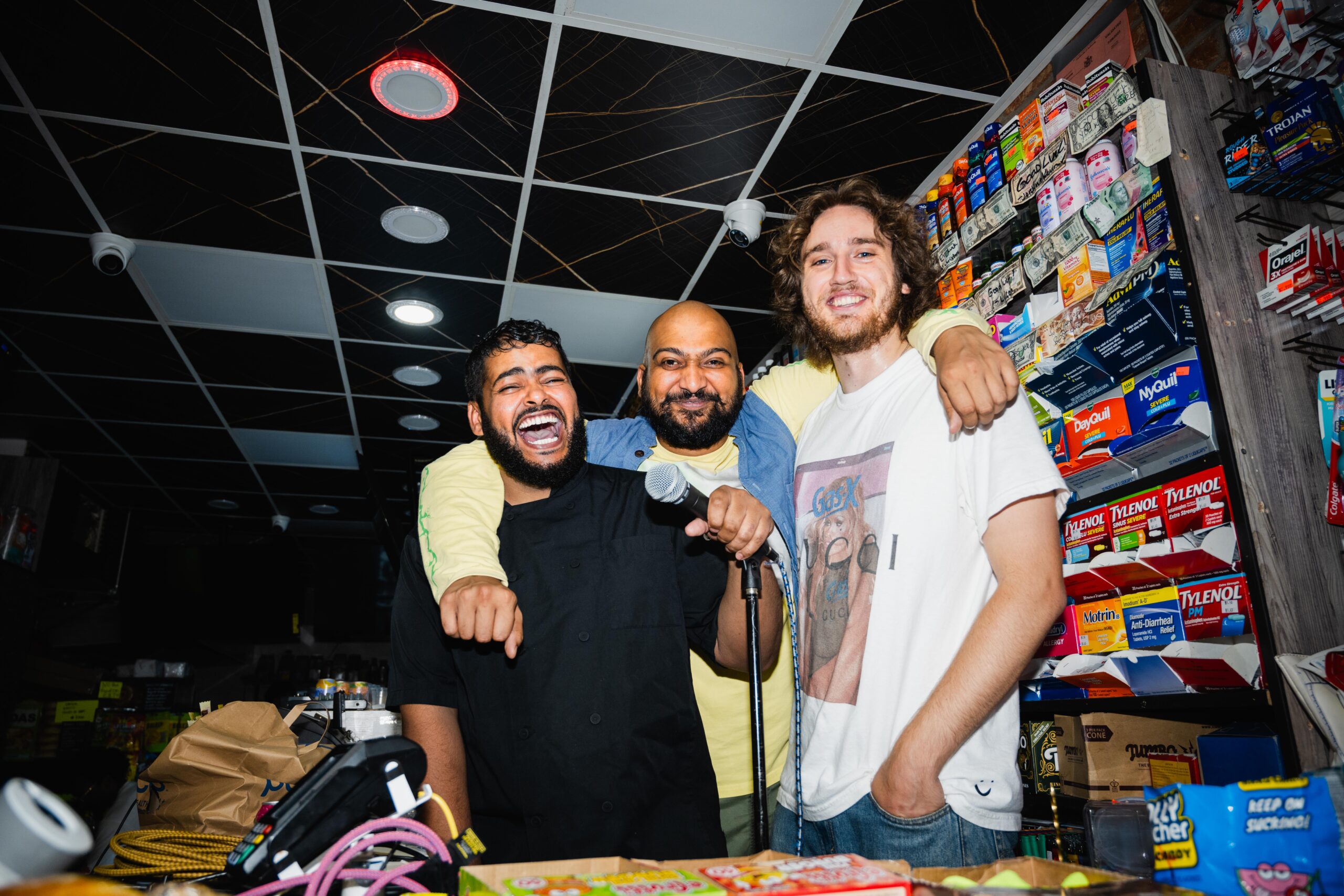 Three guys stand behind a bodega counter with a microphone