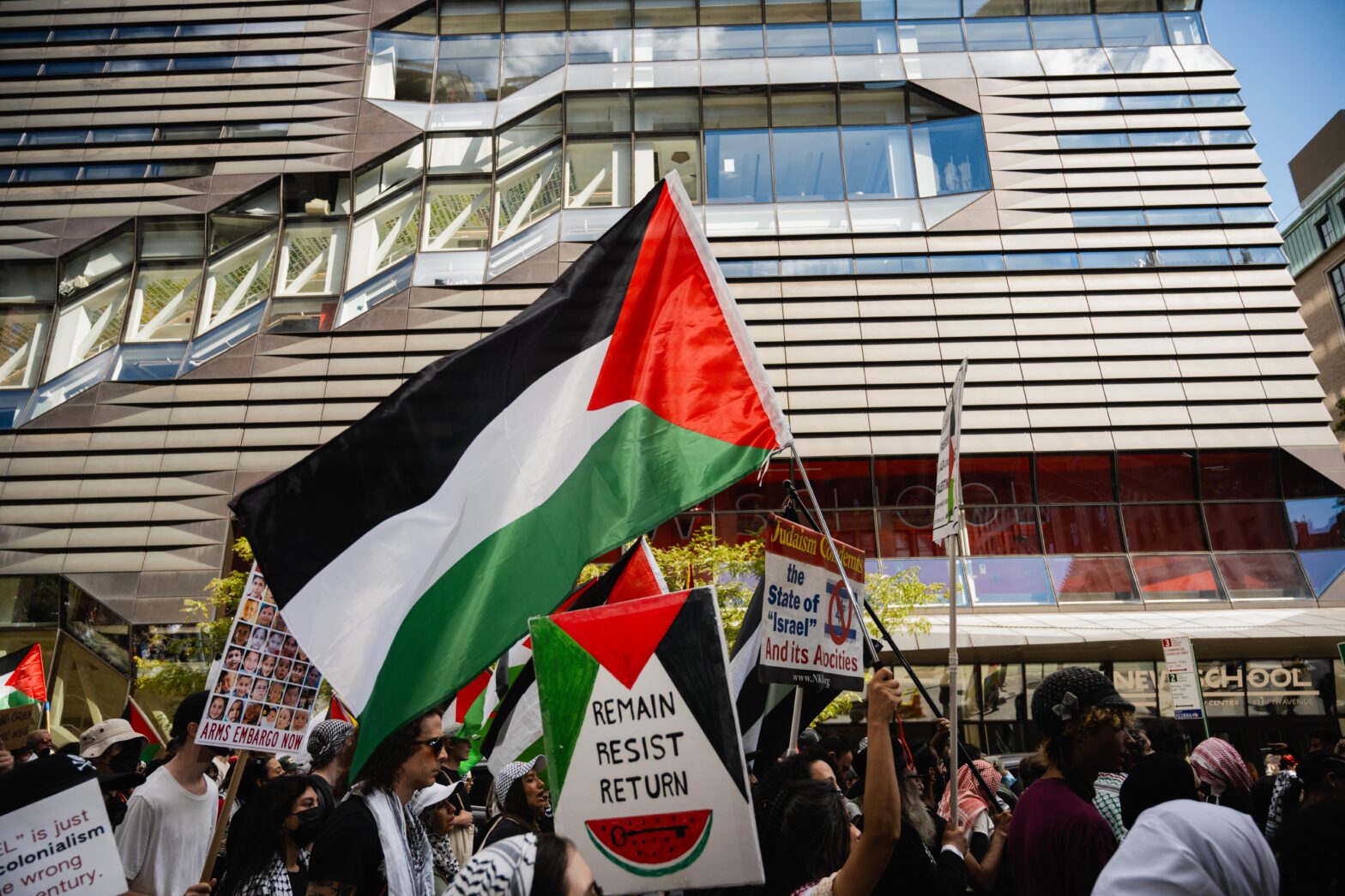 A number of protesters in front of the New School University Center entrance. In the center is a prominent Palestinian flag and below it a sign reading “Remain, Resist, Return” with the colors of the Palestinian flag and a watermelon with what appears to be a key inside.