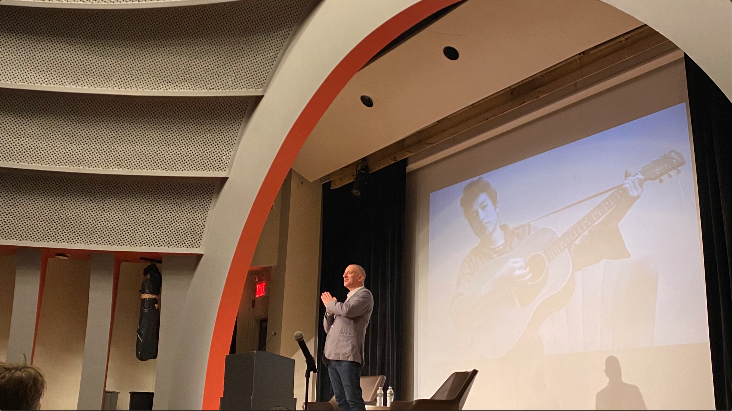 A man in a sport jacket and jeans standing on an auditorium stage in front of a screen with an image of Bob Dylan.