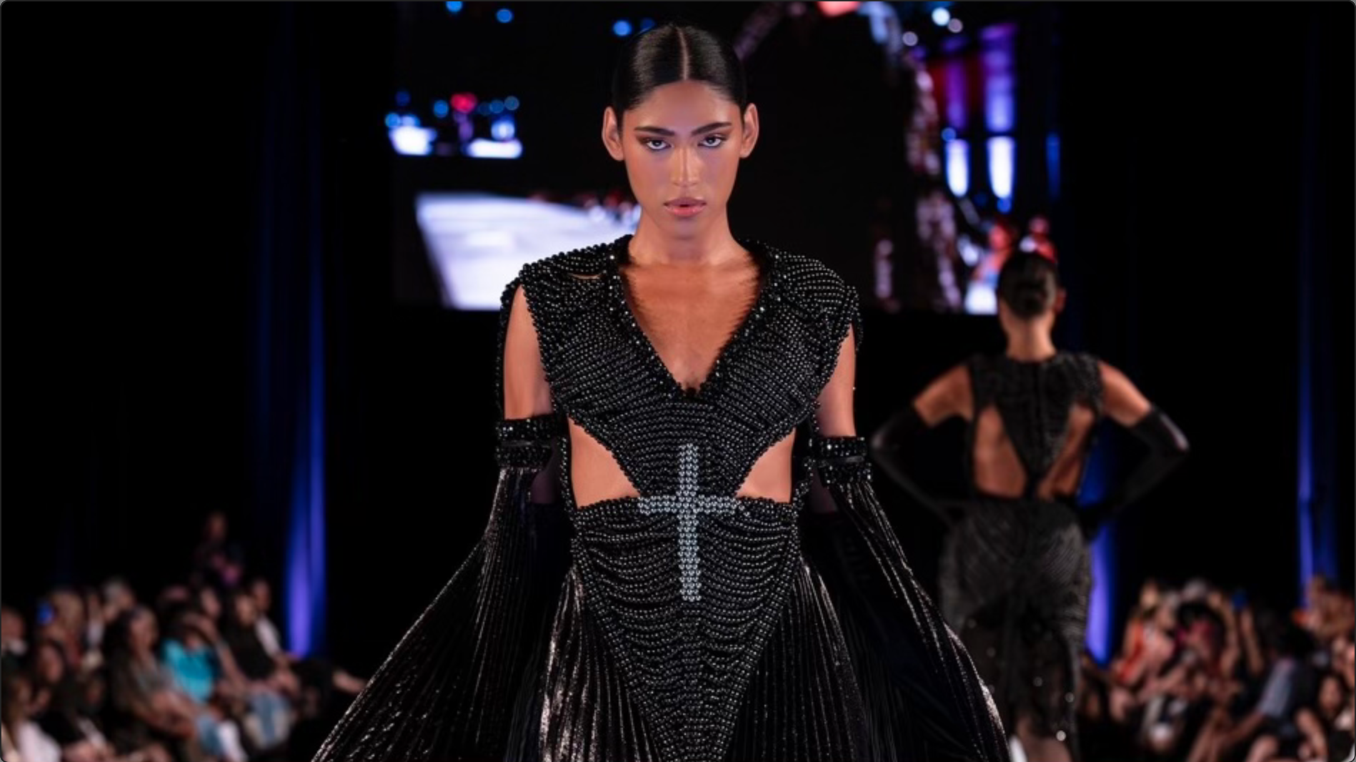 Model wears a black floor length beaded gown walking down the runway. The dress has a black pleated cape draping behind her and in the middle is a gray beaded cross.