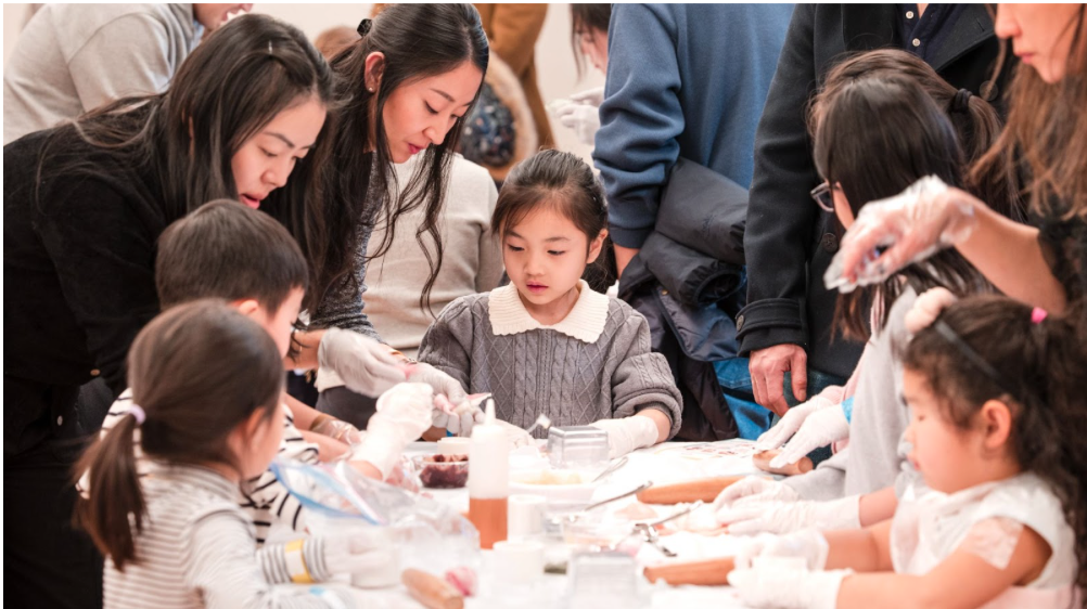 A group of children and adults gathering around the table, participating in a hands-on workshop making rice cakes.