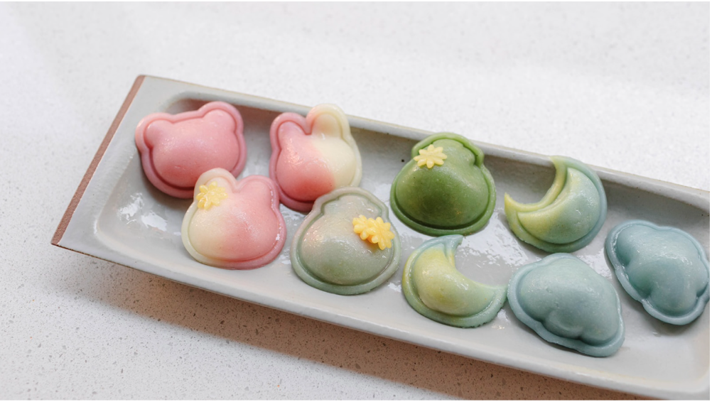 9 multicolored rice cakes shaped in animals and symbols on a grey rectangular plate. 