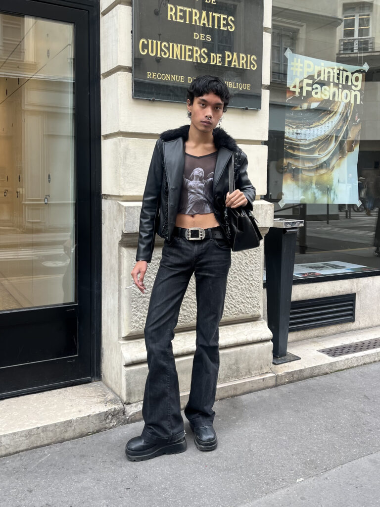 Student wearing a black leather jacket, black mesh top, black jeans, and black boots and black leather bag stands against a beige and black building in Paris, France.