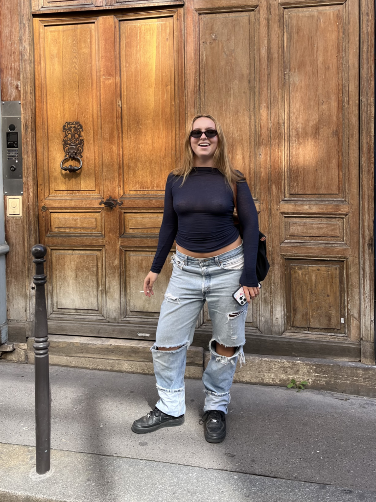 A student wearing a black long sleeve shirt, ripped blue jeans, black sunglasses, and black sneakers stands in front of an aged wooden door in Paris, France.