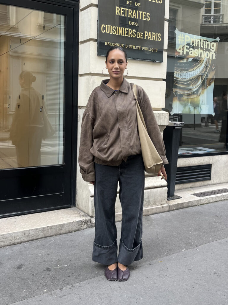 Student wearing a brown leather jacket, black jeans, beige bag, and brown tabi flats stands against a beige and black building in Paris, France.