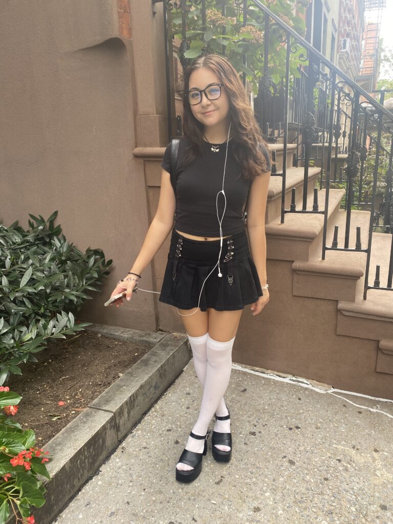 Student stands in front of the steps next to the Lang 12th Street building wearing a black crop top, a black mini skirt with buckles on the front, white knee-high socks, and black open-toed platform heels.