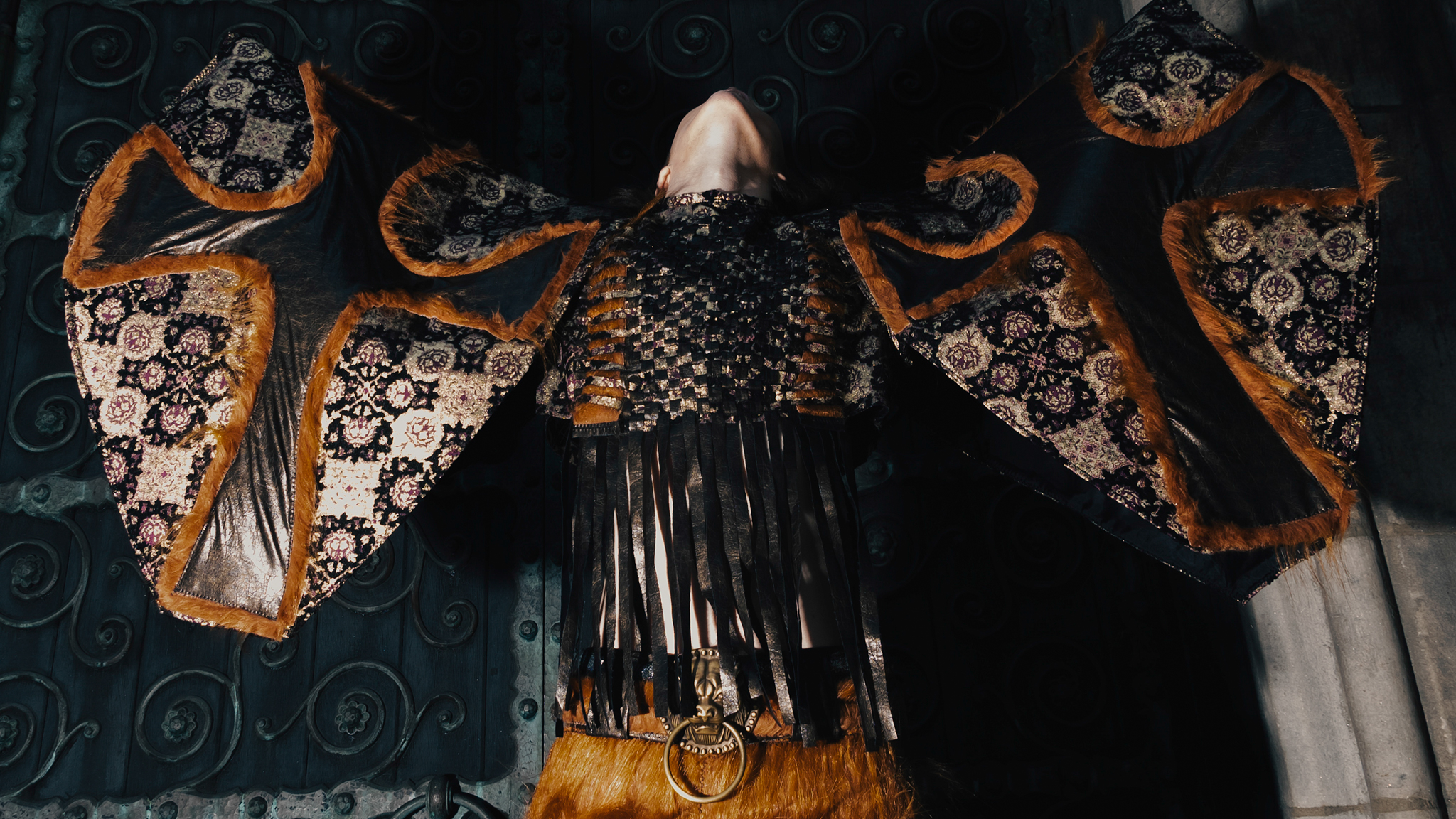 Model with arms outstretched, wearing a wing-like garment with floral tapestry, leather, and fur trim, set against a dark ornate backdrop.