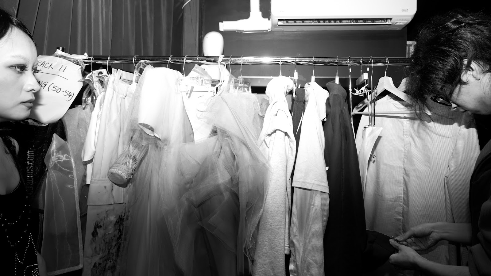 In monochromatic image, two individuals inspect garments on a labeled rack, preparing backstage for a fashion show.