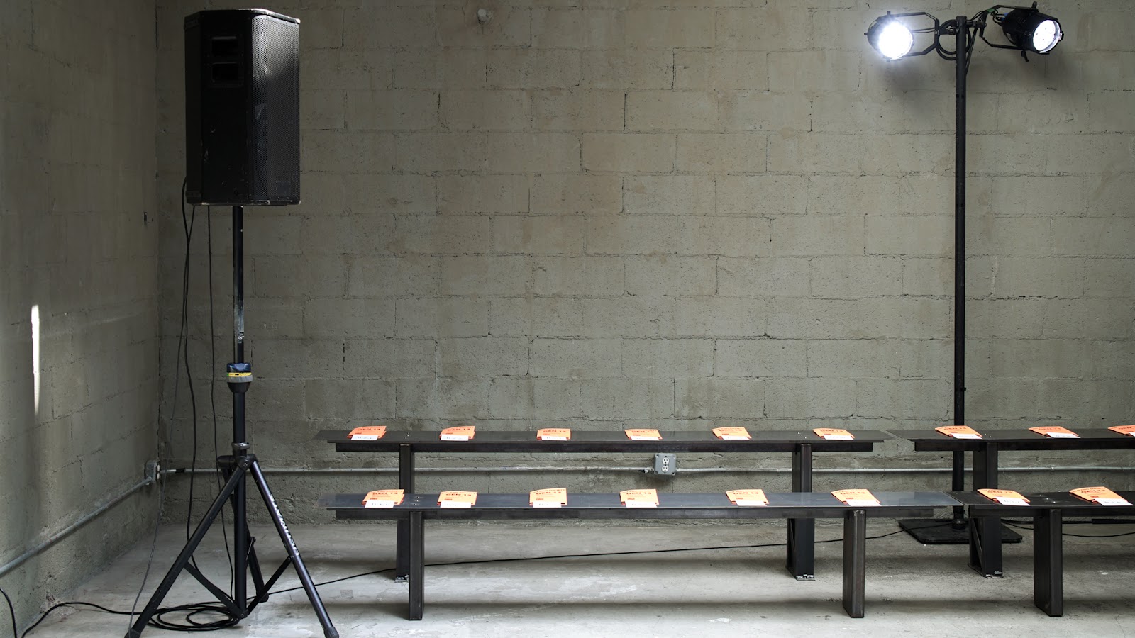 The image shows an industrial-style set up with metal benches holding orange booklets, alongside a large speaker and stage lighting against a bare concrete wall. 