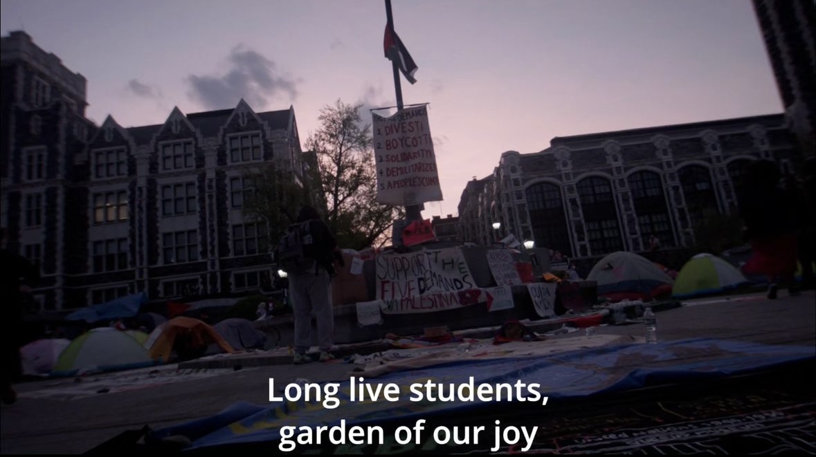 University building behind the site of a gaza solidarity encampment where tents surround a Palestinian flag on a pole, and large signs with divestment demands hang from the flag pole. the sky is pink and dark.