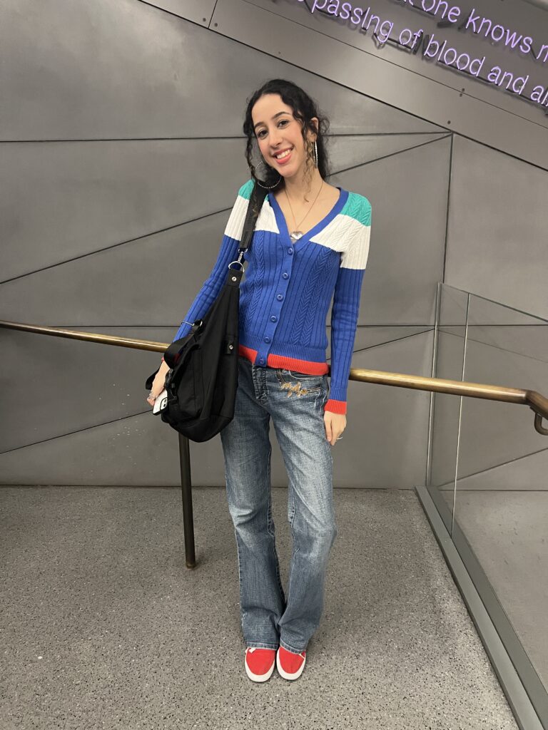 Student wears bright red sneakers, dark wash jeans with accents engraved on their left pocket, a large black sling bag, a multi-colored cardigan with a gray top buttoned underneath, and large silver hoops.