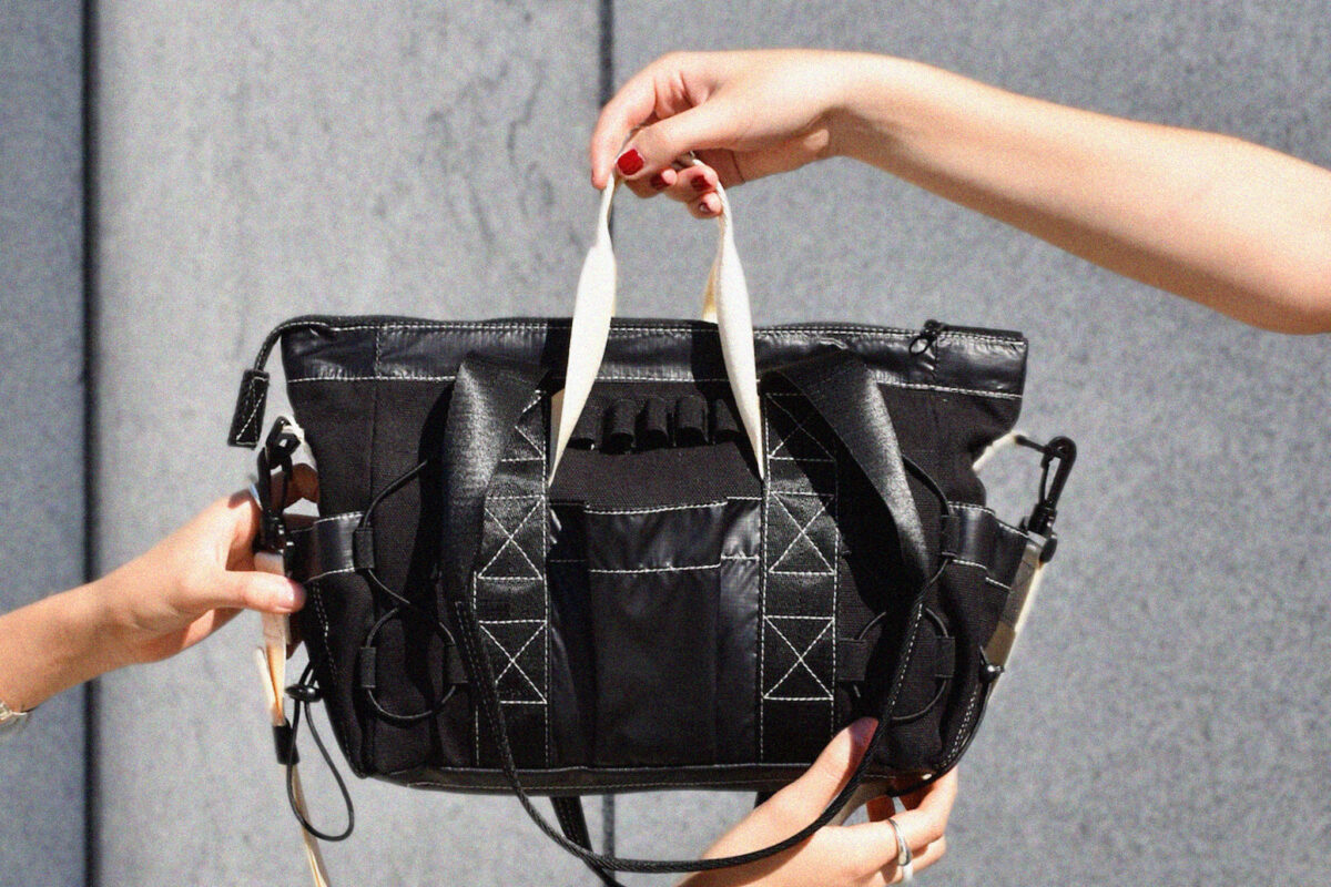 Three hands holding the small Utility Tote against a stone background. 