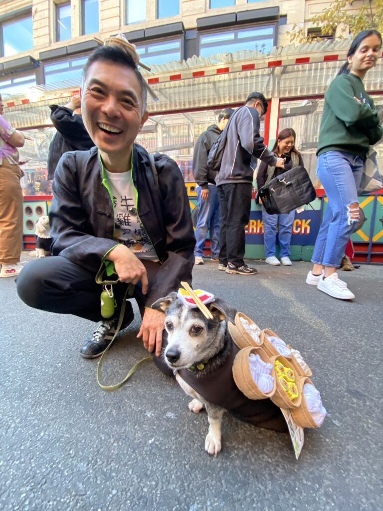 A chihuahua dachshund mix wears a tray of felt dim sum, with a sauce dish hat. In the background, his owner holds his leash and smiles at the camera. 