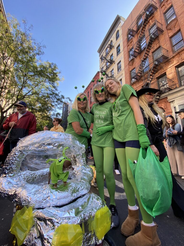 Three women dressed in all green wearing alien glasses pose for the camera in the background. In the foreground, a chihuahua in a green alien costume stands in a foil-covered stroller.