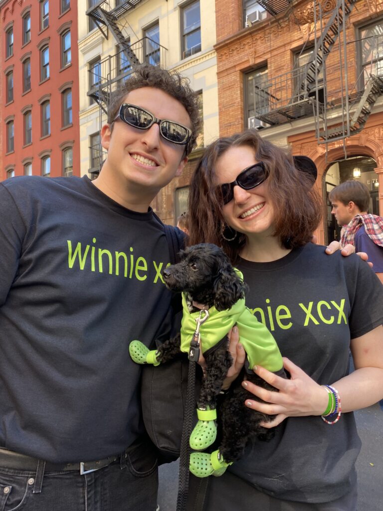 Two people wearing black shirts that read Winnie XCX in bright green font, black sunglasses, and black pants. The person on the right is holding a black toy poodle who is wearing a bright green jacket and Crocs. 