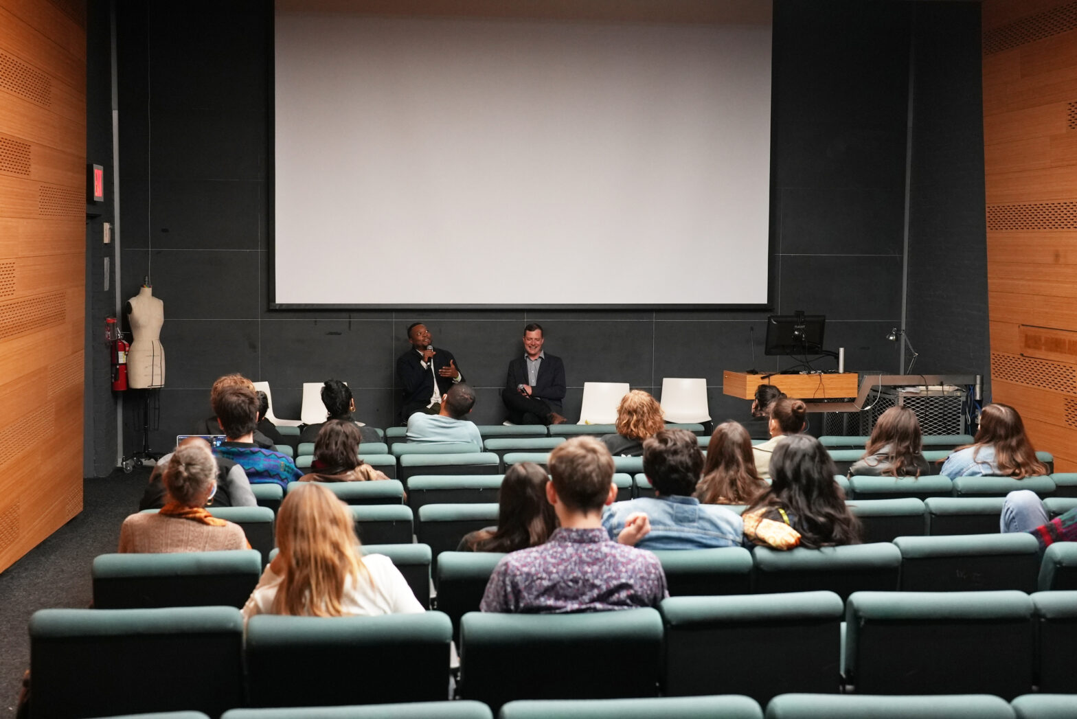 Two men sitting at the front of a small auditorium, underneath a projector screen. The audience sits in green seats in the foreground, facing away from the camera.