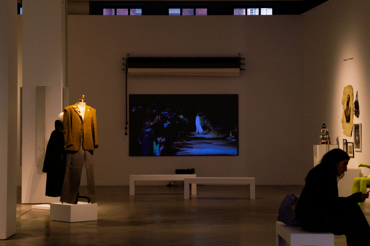 A dimly lit gallery space featuring a mannequin on the left. In the background, a video screen displays a fashion runway scene. To the right, art pieces hang on the wall. A person sits on a bench in the lower right corner. 