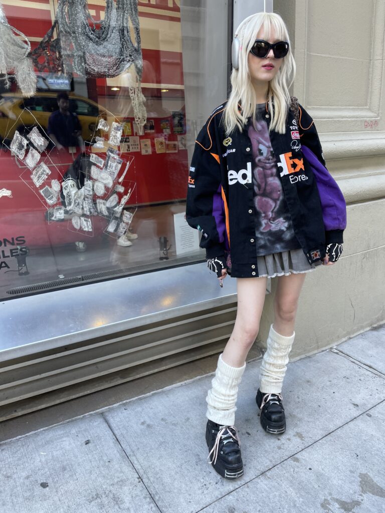 Student stands in front of the Parsons 13th Street building wearing a FedEx race car varsity jacket, a pink and black tye-dye character shirt, a pleated gray mini skirt with lace ruffle trim, white leg warmers, gray Airpods Max, large black sunglasses, and black new rock boots with pink ribbon bows. 
