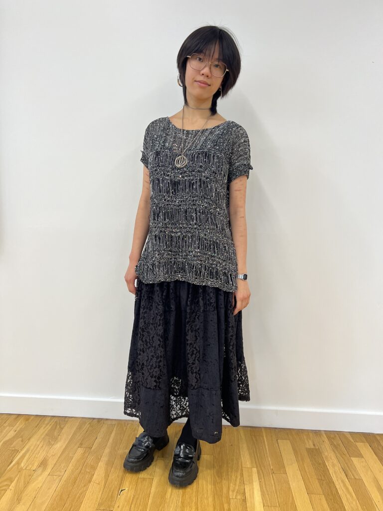 Student wears black loafers, tights, a flower-patterned lace skirt, a top underneath a gray glittery beaded garment, hexagon-shaped glasses, silver hoop earrings, a large geometric pendant necklace, and a small watch.